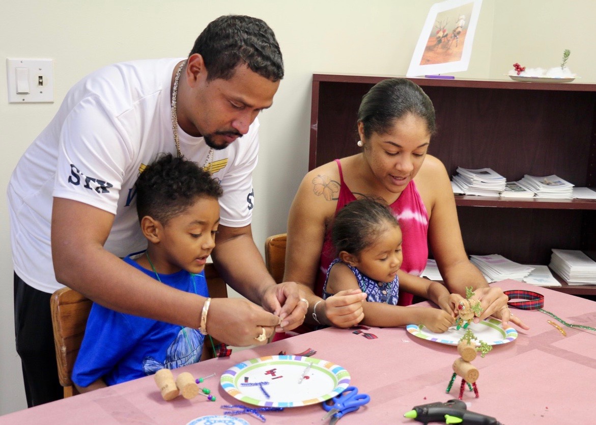 The entire Encarnacion family participated in an afternoon of creating Christmas ornaments. Christopher stands to help his son, Jahzari. Rickysha holds Baejah on her lap as they work on a reindeer. (Source photo by Linda Morland)