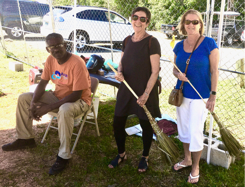 Avelino Samuel looks on while Barbie Devine and Sherri Draper wield their new brooms, woven by Edmund Roberts. (Source photo by Amy Roberts)